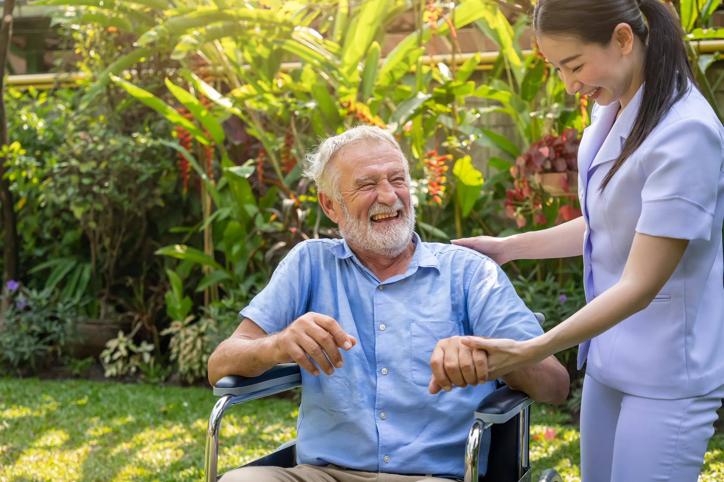 Happy nurse holding laughing elderly man hand on wheelchair in garden at nursing home Happy nurse holding laughing elderly man hand on wheelchair in garden at nursing home